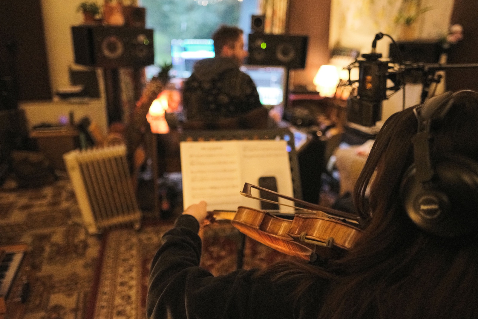 A violinist seen from behind, headphones on, playing from a printed part on a stand toward a studio microphone, the engineer at the desk beyond