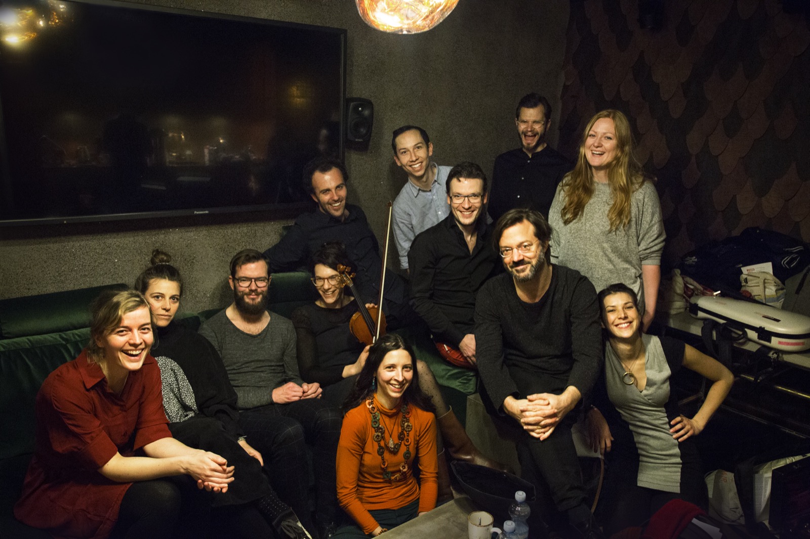 s t a r g a z e ensemble group photo — a dozen musicians on a green velvet banquette in a dim bar, a violin held up in the middle