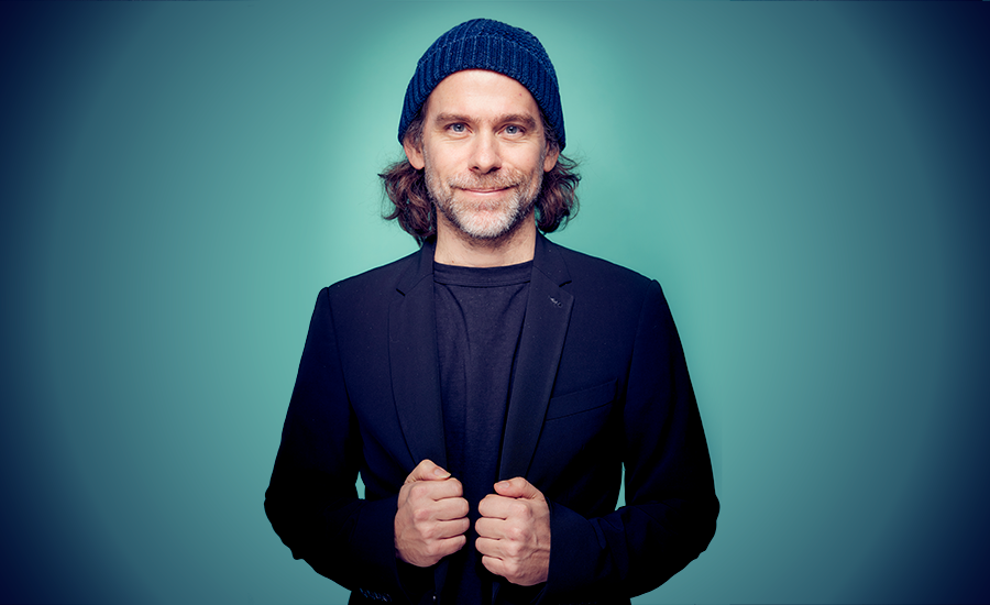 Bryce Dessner in a navy knit cap and dark jacket against a teal backdrop, smiling