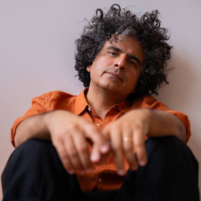 Helado Negro seated against a pale wall in an orange shirt, arms resting on his knees