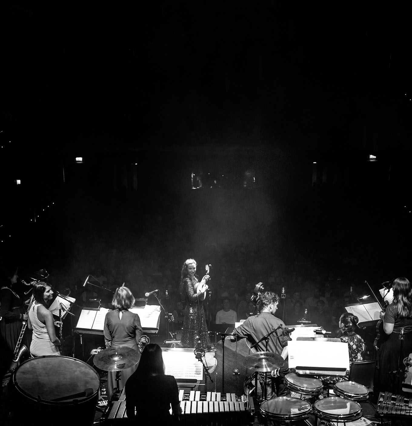 Black-and-white photo from behind the orchestra: Lisa Hannigan at center stage with a mandolin, s t a r g a z e players and music stands in the foreground, the hall dark beyond