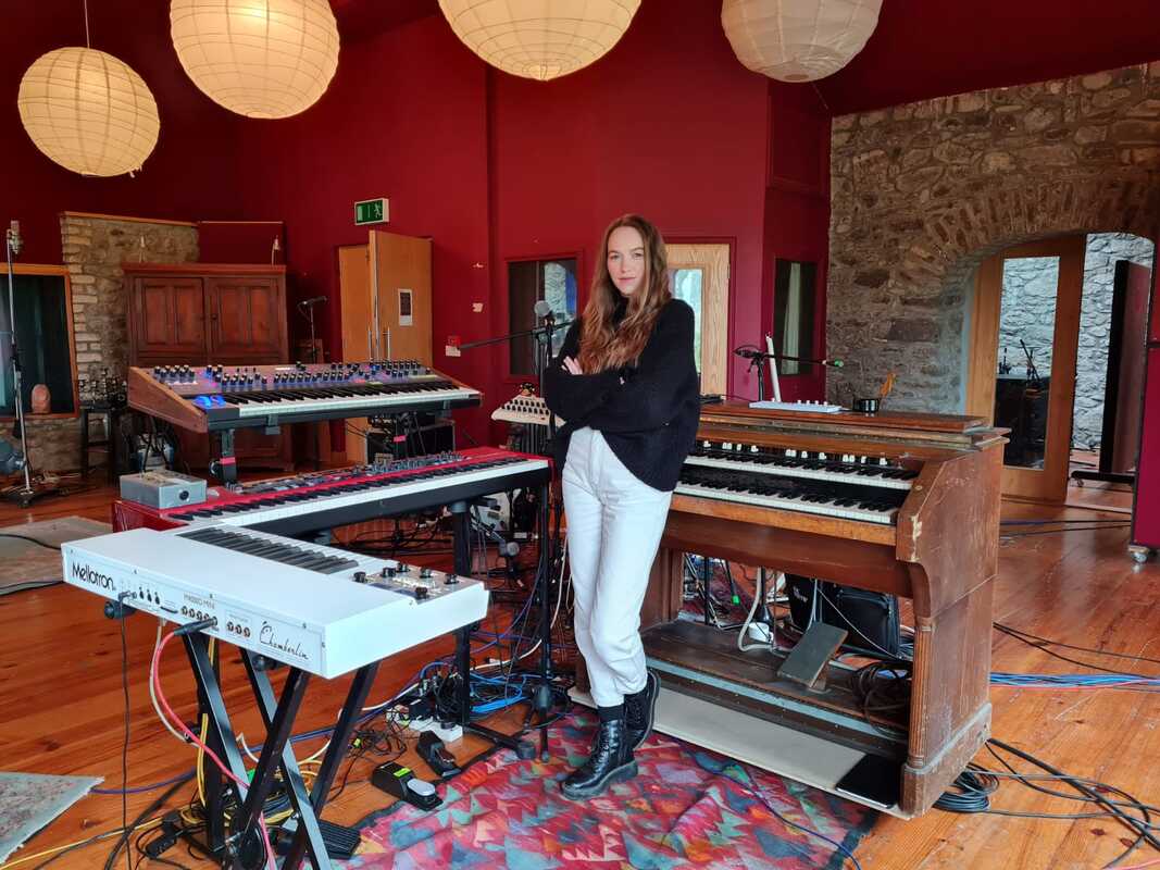 Theodora Byrne standing among a Mellotron, a Nord, and a Hammond organ in a red-walled recording studio with paper lanterns