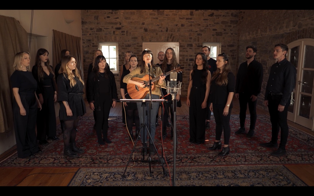 The Theodora Byrne Ensemble in person: singers in black in a stone-walled room around Maria Kelly with a guitar and a microphone