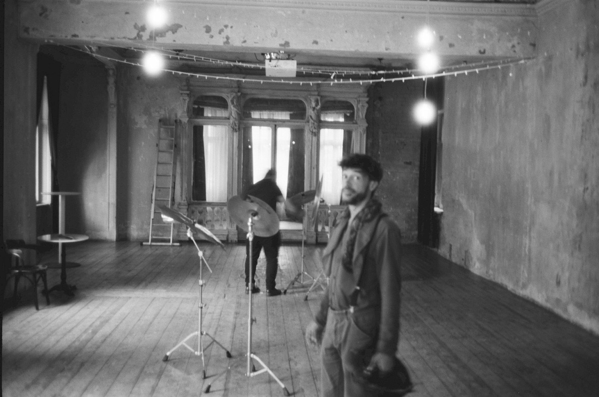 Black-and-white photo of Vincent Moon standing in a bare, peeling ballroom with a drum kit and string lights behind him